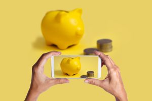 Hände nehmen Bild mit Smartphone Sparschwein und Geld auf - In der Inflation mit Finanz-Apps Geld sparen