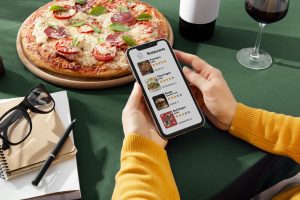 Hand hält Handy mit Restaurant-Liste, dahinter eine Pizza (Foto: Freepik) - Inflation Deutschland 2024: Unternehmen wollen Preise erhöhen