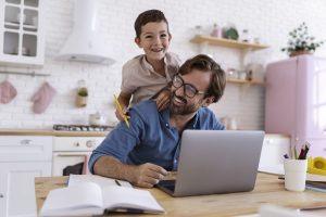 Mann arbeitet mit Laptop am Küchentisch während Kind auf ihm herumturnt (Foto: freepik) - Homeoffice oder Büro: wo die Produktivität höher ist – große Firmen-Studie