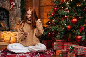 Frau im Schneidersitz vor geschmücktem Weihnachtsbaum betrachtet ihr Handy (Foto: freepik, dikushin) - Handy-Verbot an Weihnachten? So machen es Familien 2022