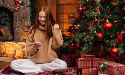 Frau im Schneidersitz vor geschmücktem Weihnachtsbaum betrachtet ihr Handy (Foto: freepik, dikushin)